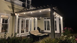 Een overdekt terras met tuinmeubilair, een pergola met lichtslingers, en een huis met grote ramen en een duidelijke deur.