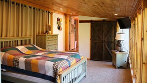 Une chambre avec un plafond en bois, un grand lit recouvert d’une couette aux couleurs vives, une commode, une lampe et un téléviseur posé sur un meuble.