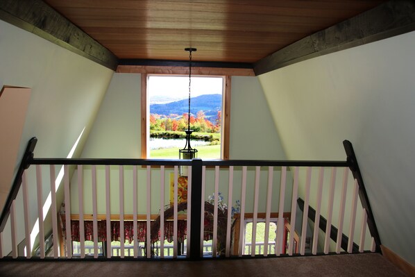 Une vue depuis un balcon doté d’une balustrade en bois, d’un luminaire suspendu et d’une fenêtre donnant sur un paysage composé d’arbres et de montagnes.