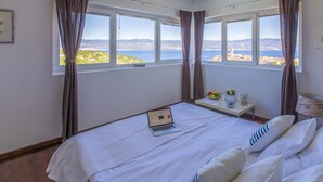 Ein Hotelzimmer mit einem Bett, einem Laptop auf dem Bett, einem kleinen Tisch mit einer Vase und einer Schale Obst, und Blick auf das Meer und die Hügel durch die Fenster.