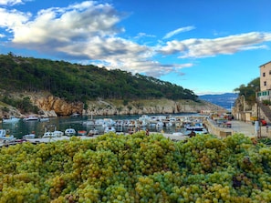 A coastal marina with boats docked, a hillside with trees, and a building with a balcony overlooking the water.