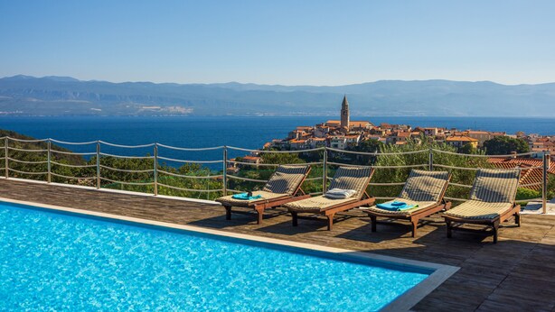 Ein Poolbereich mit Liegestühlen und Blick auf eine Küstenstadt und Berge.