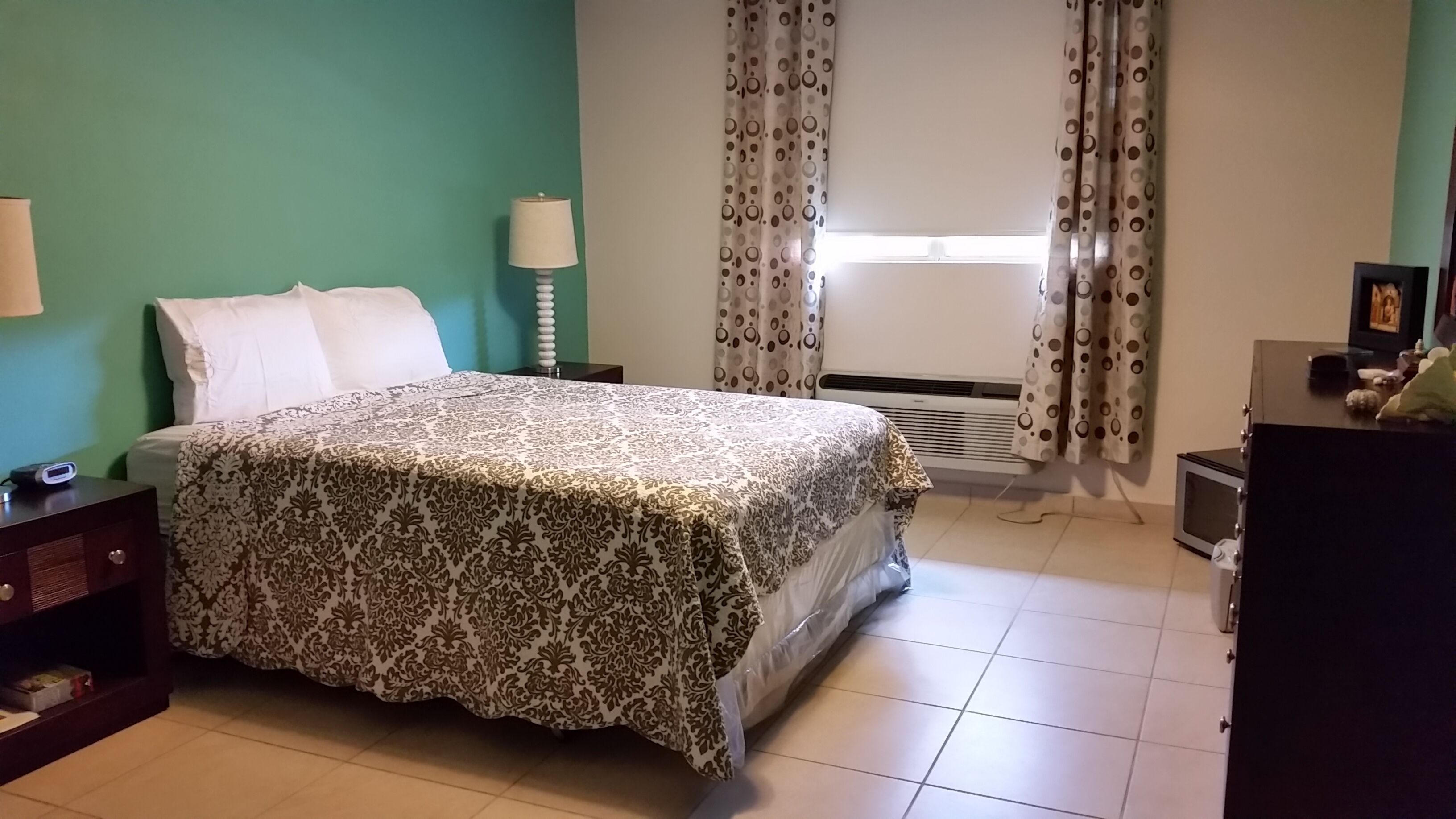 Une chambre à coucher comprenant un lit, une table de chevet, une lampe, une fenêtre avec des rideaux, un climatiseur et une commode.