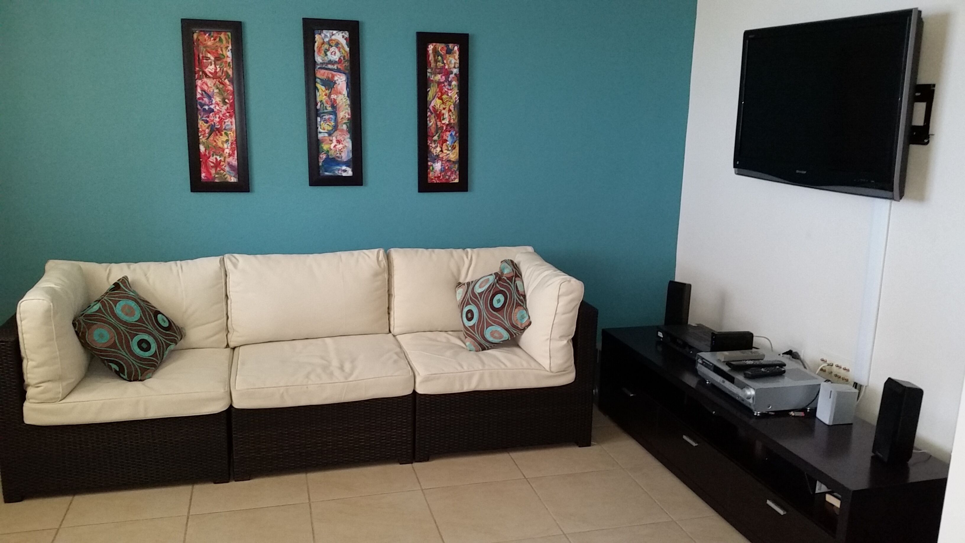 Una sala de estar con un sofá color beige, dos almohadones decorativos, un televisor de pantalla plana y un mueble de entretenimiento negro con dispositivos electrónicos.