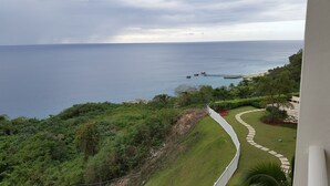 A coastal view from a balcony with a grassy area, a white fence, and a pier in the distance. - Beautiful Mediterranean Style Ocean View Villa, Spectacular Sunsets!  (Aguadilla)