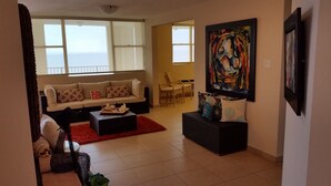 A living room with a sofa, coffee table, and chairs. There is a large painting on the wall and a window with blinds. - Beautiful Mediterranean Style Ocean View Villa, Spectacular Sunsets!  (Aguadilla)