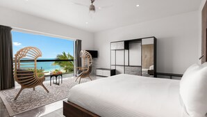 A modern hotel room with a large bed, two chairs, a TV, and a balcony with a view of the ocean.