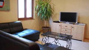 Een woonkamer met een zwarte leren bank, een glazen salontafel, een potplant, een televisie op een houten kast en een raam met jaloezieën.