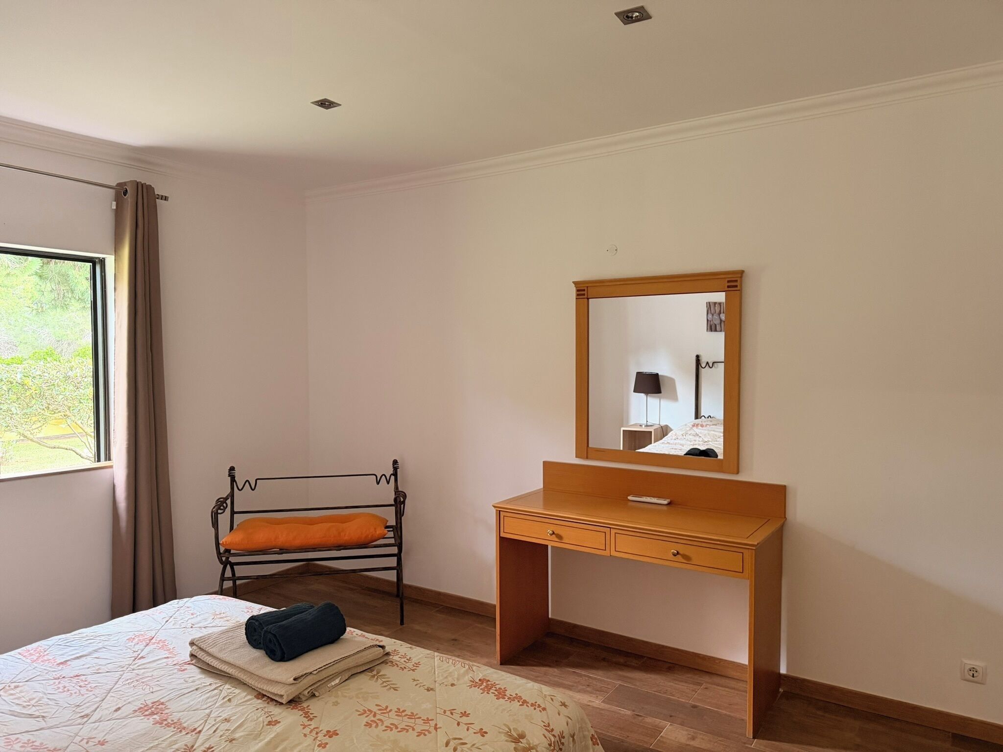 Une chambre à coucher avec un lit, un bureau en bois, un miroir, une fenêtre avec des rideaux et un radiateur.