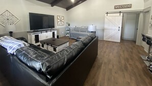 A spacious living room with a black leather sofa, a television, and a 'welcome' sign. - Custom Barndominium Perfect For Romantic Getaways, Lake or Family Vacations (Caldwell)