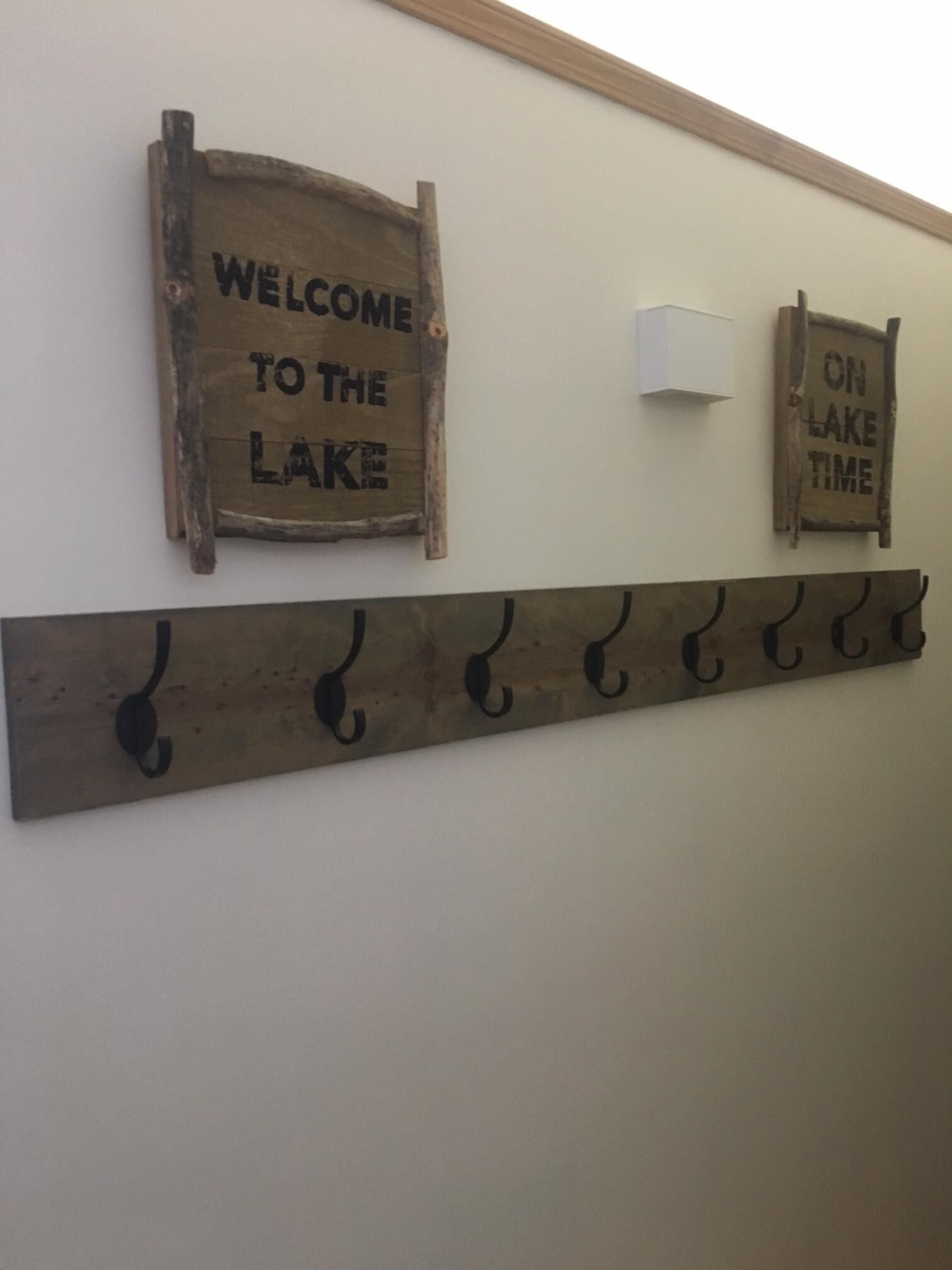 Two wooden signs on a wall, one reads "WELCOME TO THE LAKE" and the other "ON LAKE TIME". Below, there is a metal rack with hooks.