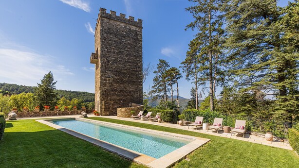 Una piscina con sillas de descanso, una torre de piedra y montañas al fondo.