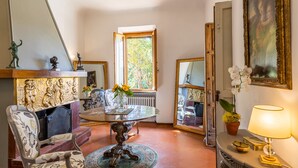Una habitación con una mesa redonda, sillas, un radiador, una ventana que da a árboles y una lámpara.