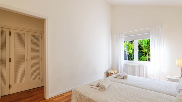 Quarto com duas camas, uma janela com cortinas, piso de madeira e uma porta com venezianas.