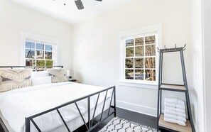 Un dormitorio con una cama, dos ventanas, una mesita de noche, un espejo y un estante con toallas.