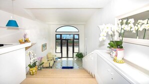 Un pasillo luminoso con una escalera que conduce a una puerta con paneles de vidrio. Hay orquídeas en macetas a la izquierda y un espejo a la derecha.