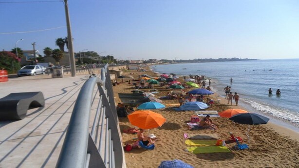 Ein Strand mit badenden Menschen, Sonnenschirmen und einem Metallgeländer entlang der Küste.