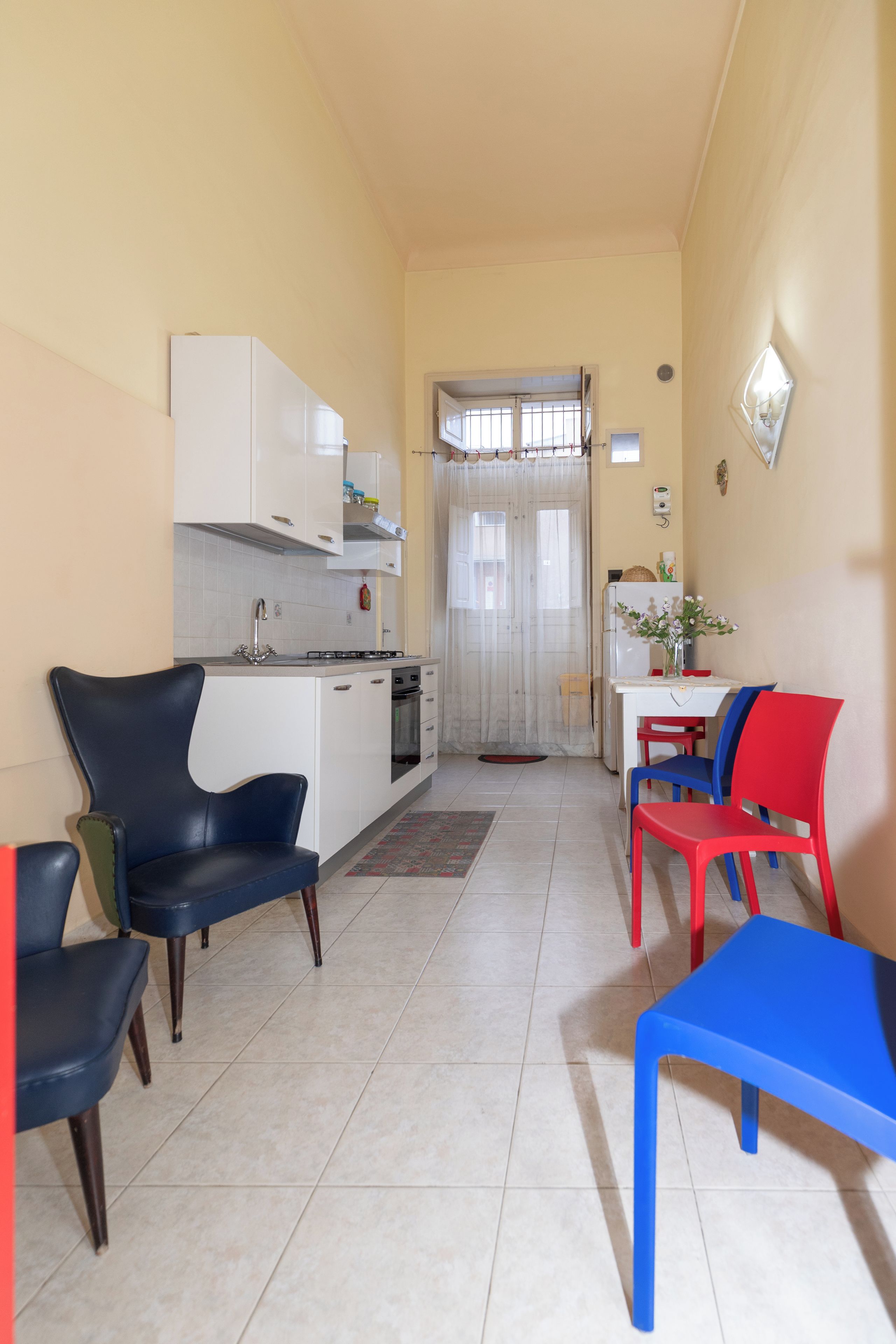 Una cucina compatta con mobili bianchi, un fornello a gas e un lavandino. Sono presenti due sedie e un tavolo con un vaso di fiori.