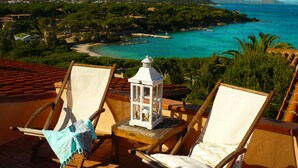 Una terraza con dos sillas de madera y una pequeña mesa, con vistas a un paisaje costero que incluye una playa y vegetación.