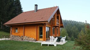 Een houten hut met een schuin dak, een schoorsteen en een klein terras met buitenmeubels.