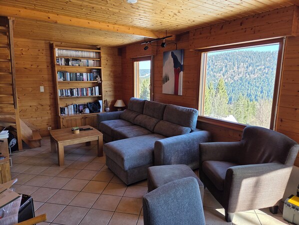 Interior acogedor de una cabaña con sofá modular, estantería de madera y un ventanal con vistas al bosque.