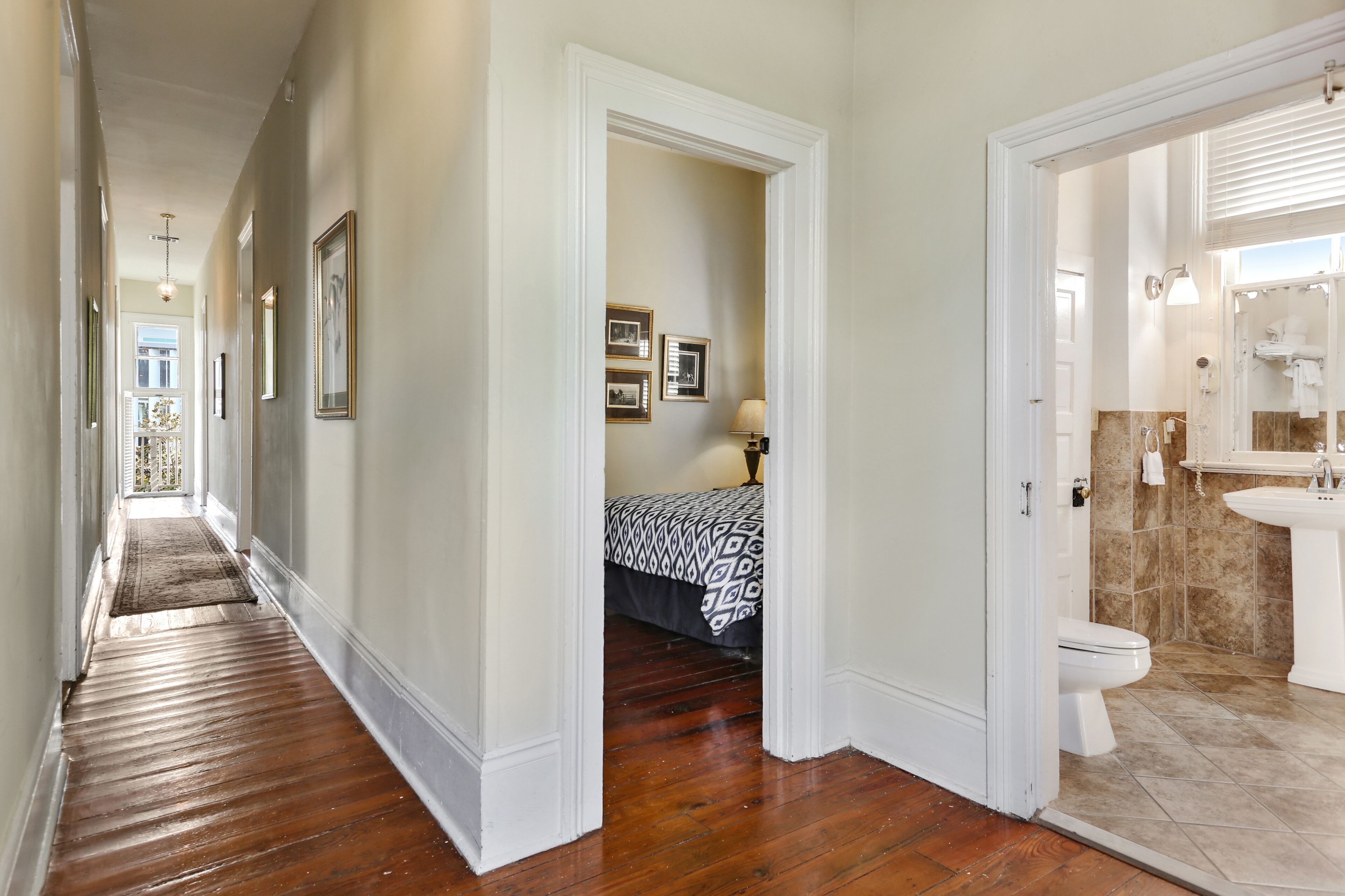 A hallway with wooden flooring, a bed with patterned bedding, and a bathroom with a sink and toilet.