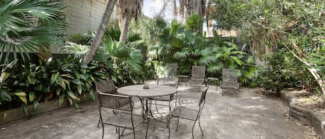 A patio with a round table and metal chairs, surrounded by lush greenery and a concrete floor.
