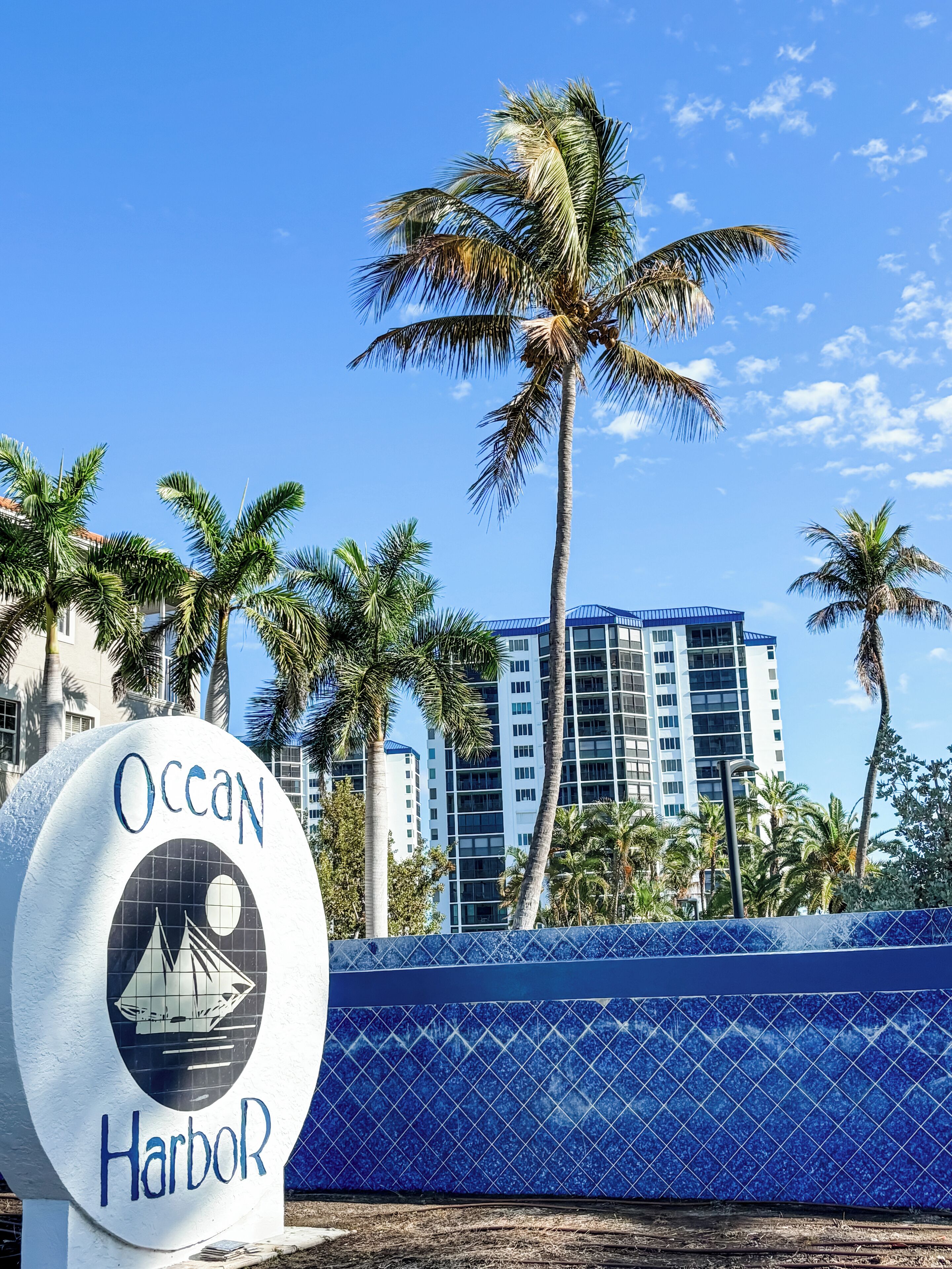 A sign for Ocean Harbor with a sailboat graphic, palm trees, and a modern building in the background.