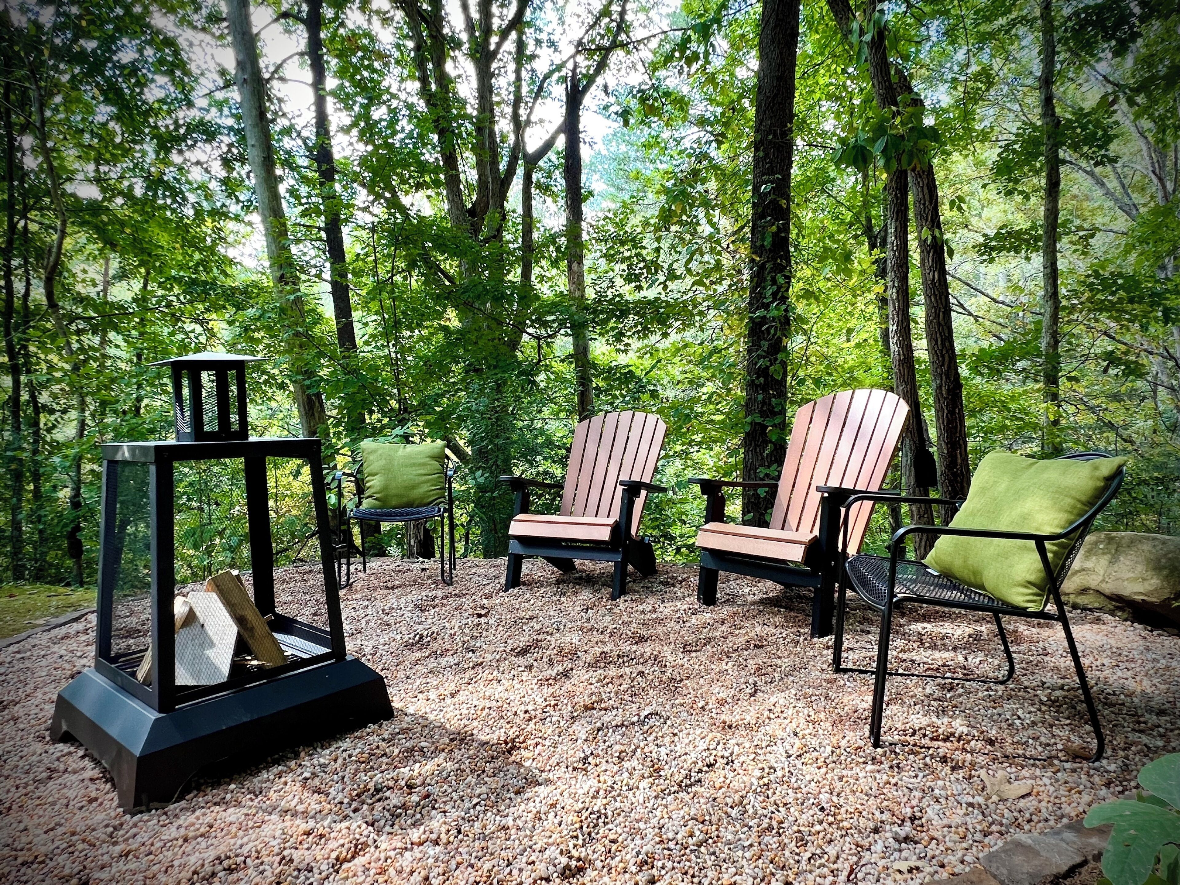 A forested area with Adirondack chairs and a fire pit.
