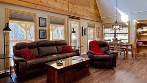 Una sala de estar acogedora con techo alto, vigas de madera y ventanas amplias. Hay dos sofás de cuero con cojines rojos, una mesa de centro de madera y un comedor con sillas de madera y una araña.