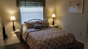 A bedroom with a bed, two bedside lamps, a window with blinds, and a framed picture on the wall.