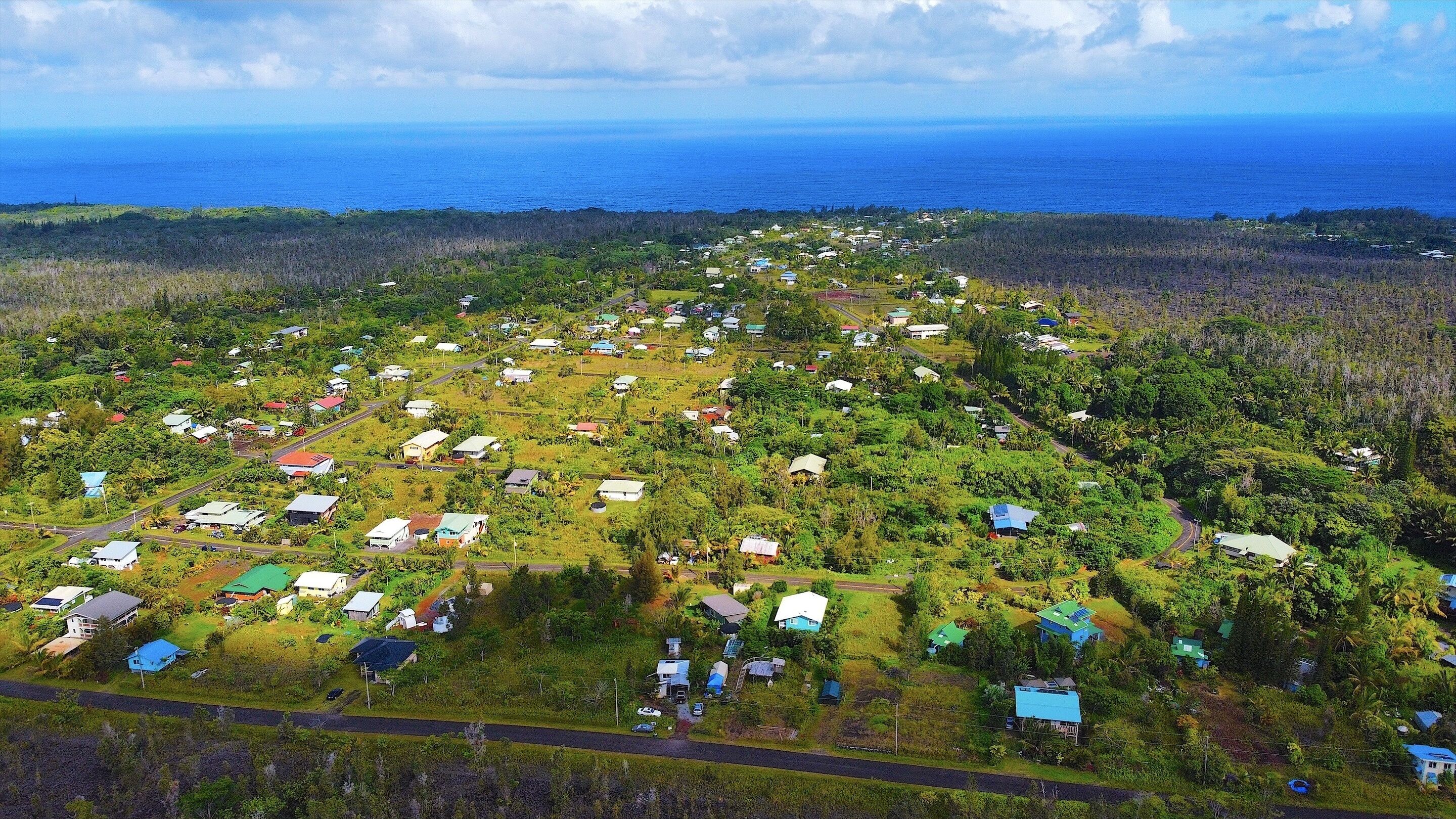 An aerial view of a coastal village with houses scattered across a hilly terrain, roads connecting the area, and the ocean in the background.