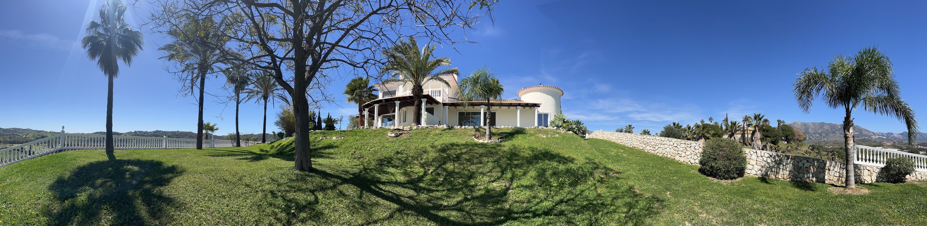 Una casa grande de dos pisos con fachada blanca y una torre circular, rodeada por un muro de piedra y palmeras.