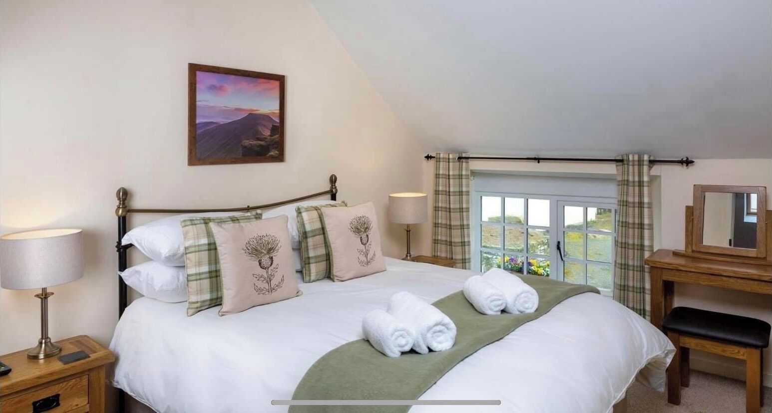 A neatly made bed with a green throw, a bedside lamp, and a painting of a landscape on the wall.