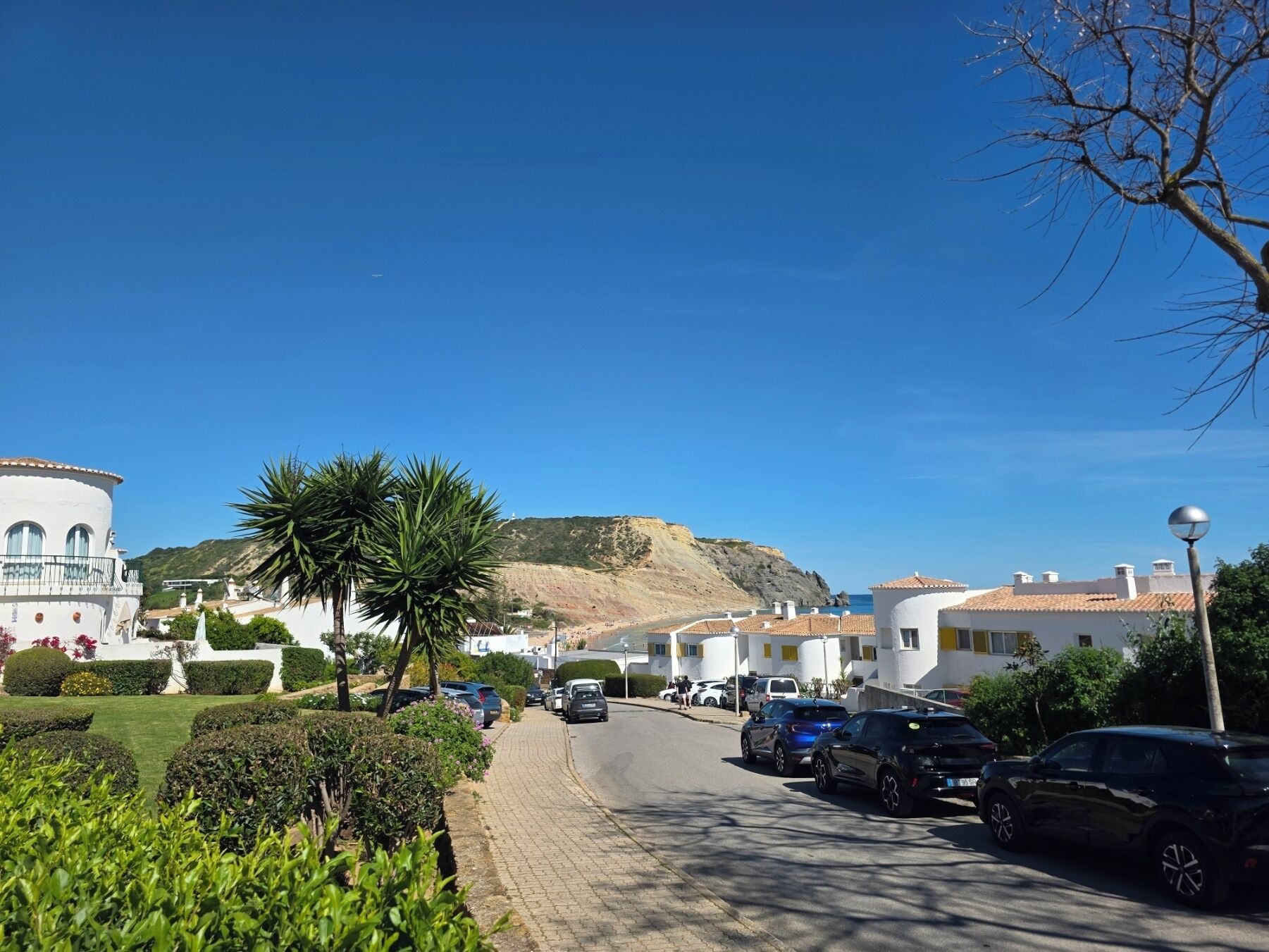 A coastal street with parked cars, white buildings with balconies, and a hill in the background.