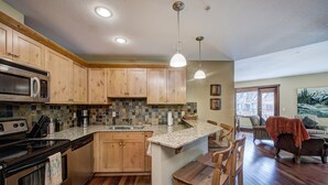 A kitchen with wooden cabinets, a built-in microwave, a stove, and a sink. There is a dining area with a table and chairs. - Canmore Luxury Condo - Panoramic Mountain Views - Prime Downtown Location (Canmore)