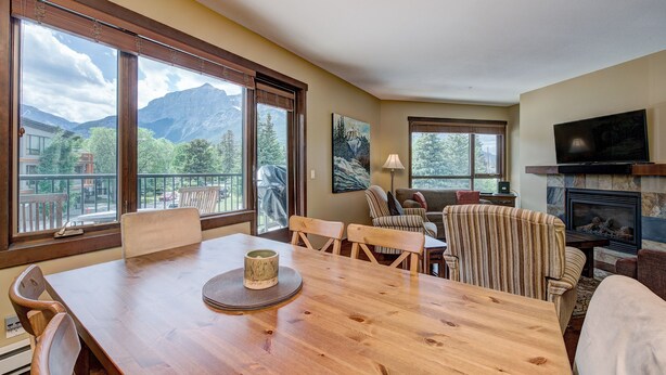 A dining area with a wooden table, chairs, and a view of mountains through large windows. - Canmore Luxury Condo - Panoramic Mountain Views - Prime Downtown Location (Canmore)