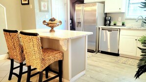 Una cocina con una isla central, dos sillas de mimbre, un refrigerador de acero inoxidable y un lavavajillas.