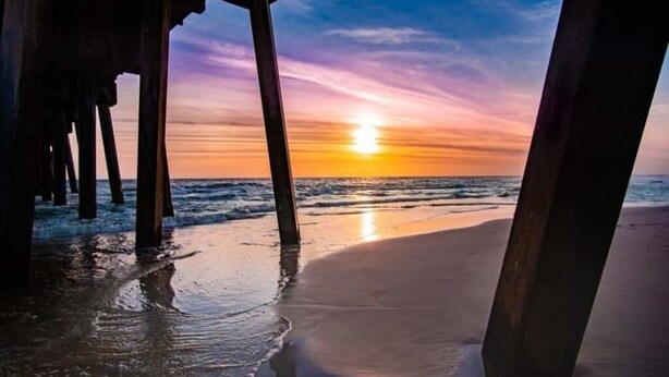 Escena de playa con un atardecer, que presenta estructuras de madera que podrían ser parte de un muelle o un edificio frente al mar.