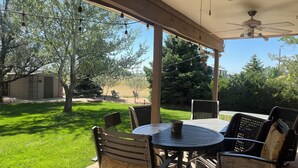 An outdoor patio with a round table and chairs, a ceiling fan, and a view of a grassy yard and trees. - 5-bedroom vacation home Mead,Co. /Easily access I-25 DenverBoulderFtCollinsEstes (Mead)