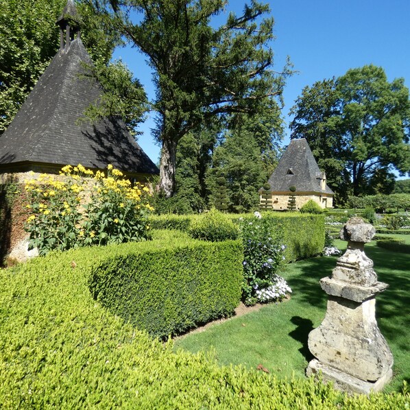 Ein Garten mit zwei Steinhäusern, eines mit einem Strohdach und das andere mit einem Schieferdach, umgeben von gepflegten Hecken und blühenden Pflanzen.