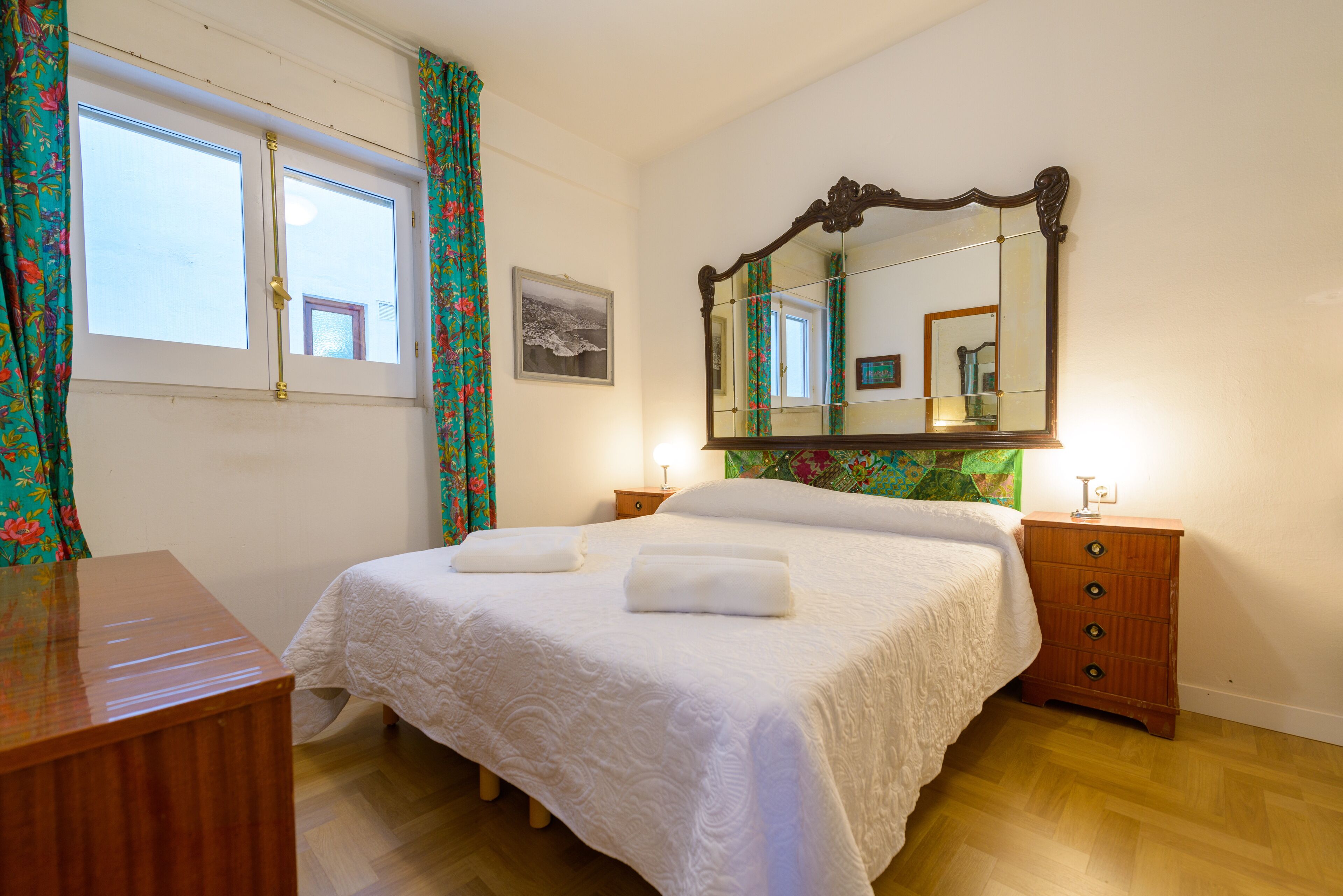 A neatly made bed with white linens, a wooden dresser, a nightstand with a lamp, and a large mirror above the dresser.