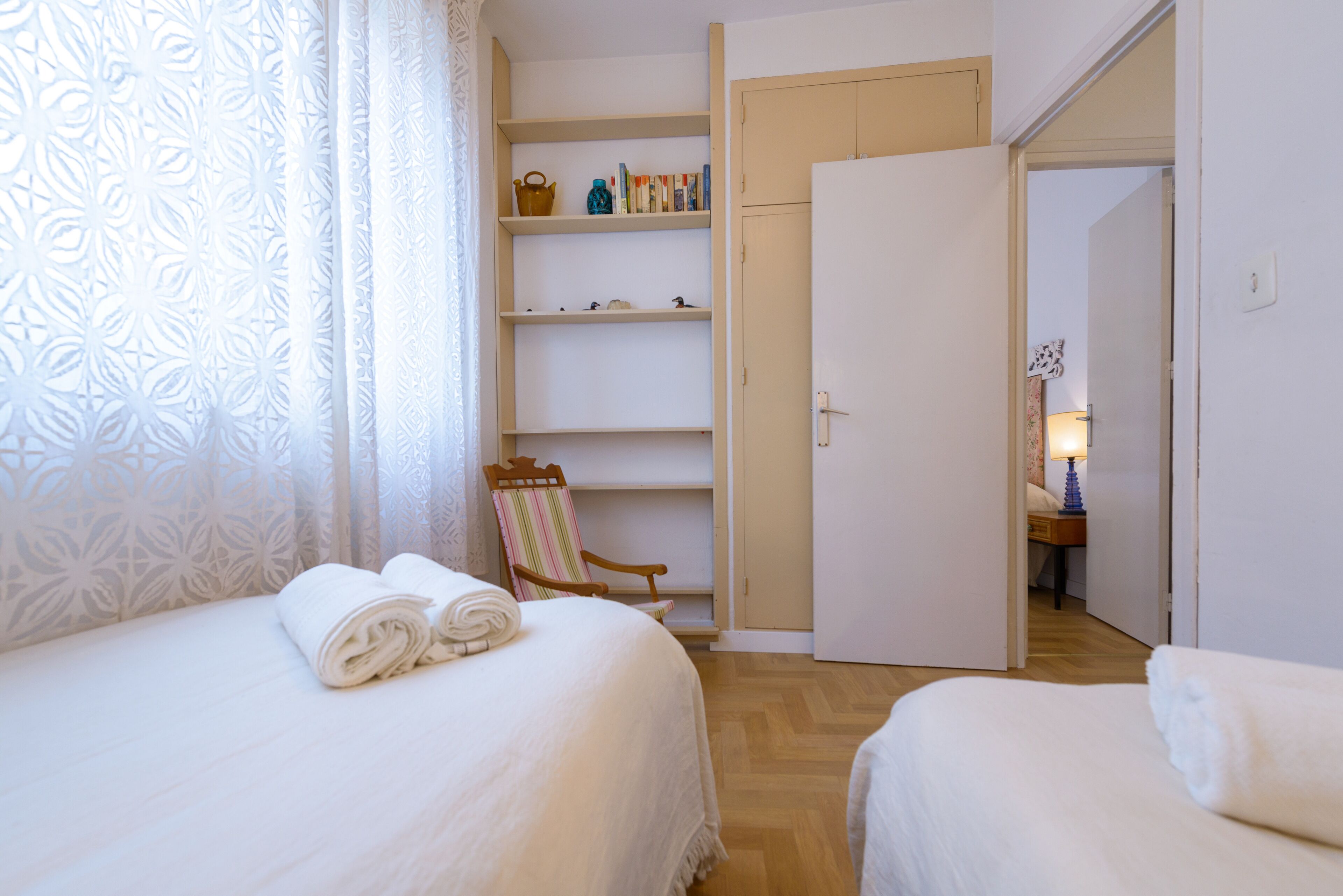 Quarto de hotel com duas camas, uma cadeira de madeira, uma estante e uma porta que dá para outro cômodo.