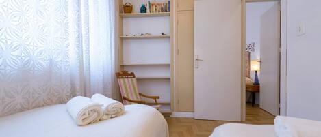 Habitación de hotel con dos camas, una silla de madera, una estantería y una puerta que da a otra habitación.
