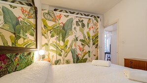 Een hotelkamer met een groot bed, een nachtkastje met een lamp, een houten dressoir en een muur met muurschilderingen van tropische planten en bloemen.