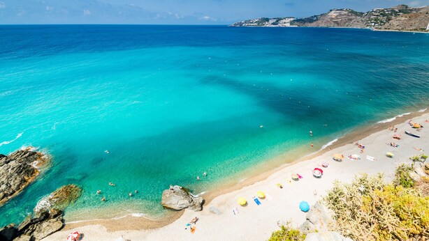 Een strand met helder turquoise water, een zandstrand en enkele parasols.