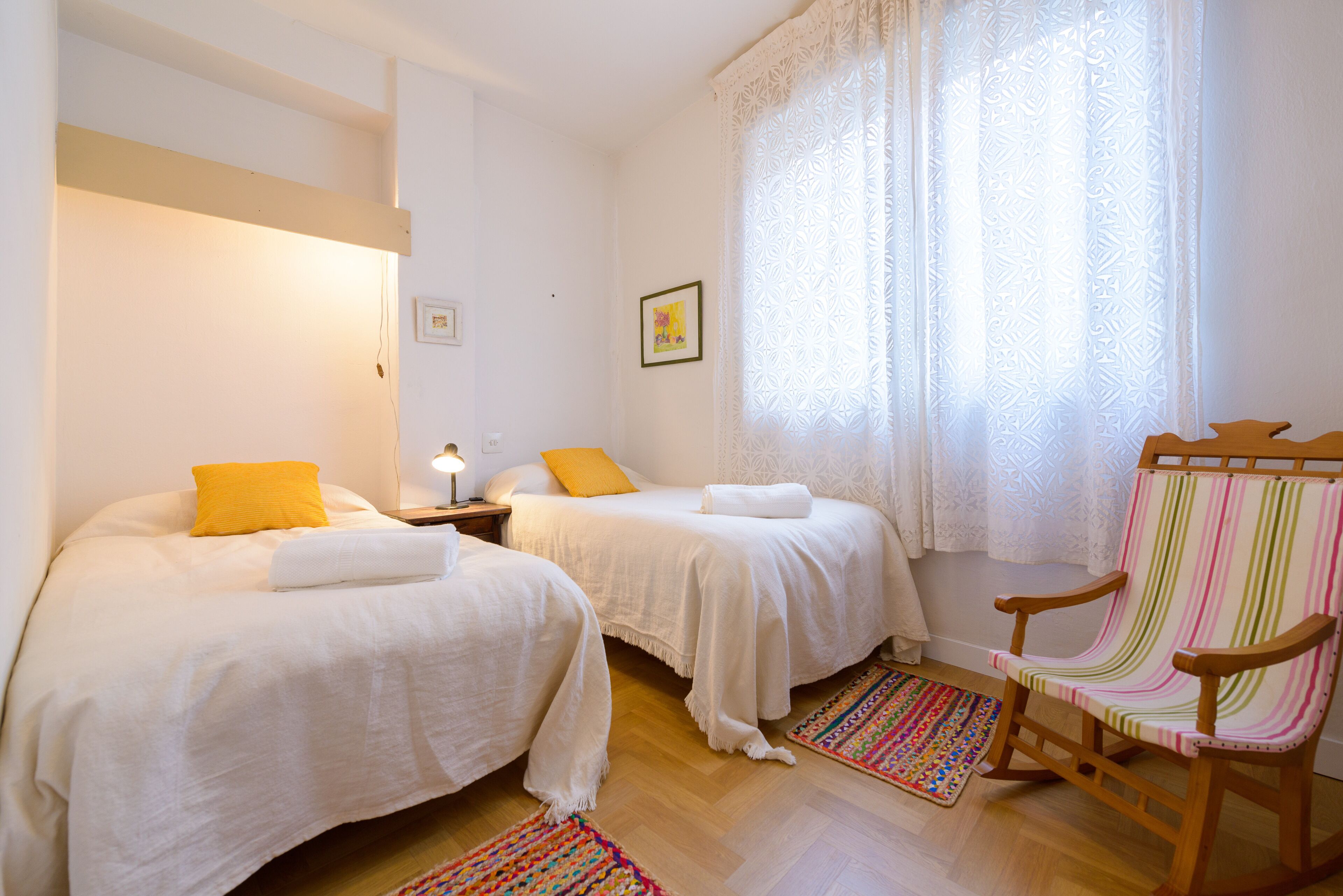 A bedroom with two beds, a wooden rocking chair, a colorful rug, and a window with lace curtains.