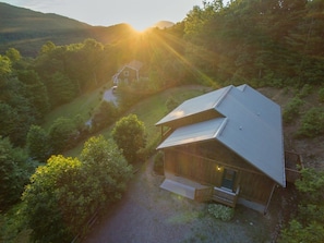 A wooden cabin with a sloped roof, surrounded by dense forest, and a clear path leading to it. - Hot Tub+Fire Pit, Grill, Fireplace, WiFi, Private! (Burnsville)