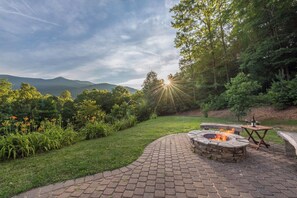 A fire pit area with a brick pathway, surrounded by lush greenery and a view of distant mountains. - Hot Tub+Fire Pit, Grill, Fireplace, WiFi, Private! (Burnsville)