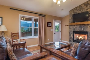A cozy living room with a stone fireplace, leather sofas, a glass coffee table, and a large window with curtains. - Sunset Ridge! Hot tub+Fire Pit+Grill, WiFi, Mtn Views, Easy Access, Nearby Town! (Burnsville)